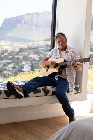 Mature man playing acoustic guitar on window bench with scenic valley and mountain view