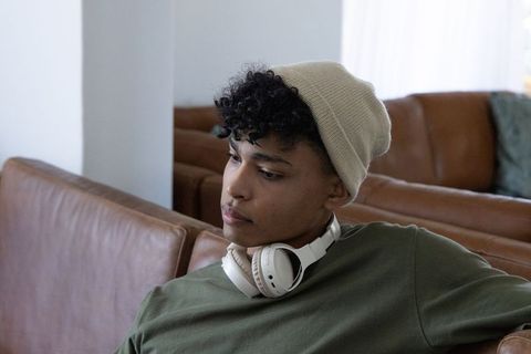 Thoughtful african american man sitting on leather sofa wearing beanie and headphones