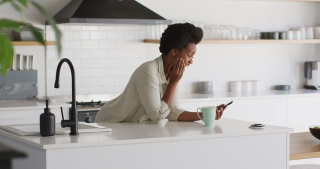 Happy african american woman drinking coffee and talking on smartphone smartphone in kitchen. domestic lifestyle, spending free time at home.
