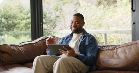 African american man relaxing on leather couch using tablet and smiling near window