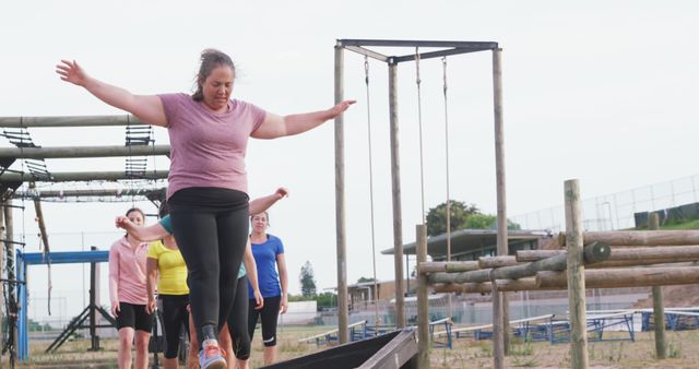 Woman participates in an outdoor fitness class, balancing on an obstacle course beam while fellow participants follow. Ideal for promoting fitness programs, exercise routines, community activities, and teamwork-focused events.