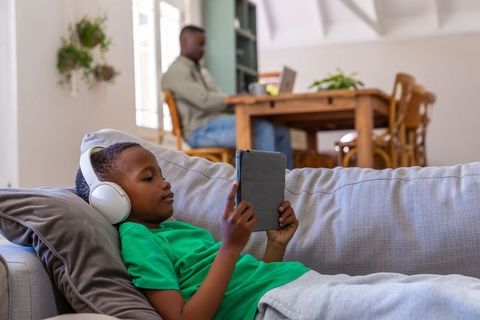 African american boy relaxing with tablet while father working remotely at dining table