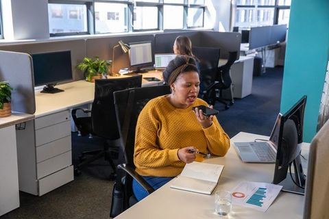 African American woman recording voice memo at desk in modern open-plan office with laptop