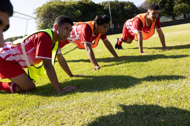 Multiracial soccer team players wearing red jersey and sports bibs doing push-ups on grassy land. Playground, summer, unaltered, soccer, sport, teamwork, competition, exercising and training.