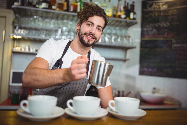 Young male barista smiling while preparing coffee at a cafe counter. Ideal for use in articles or advertisements related to coffee shops, hospitality industry, customer service, and small businesses. Can be used to depict friendly service and professional barista skills.