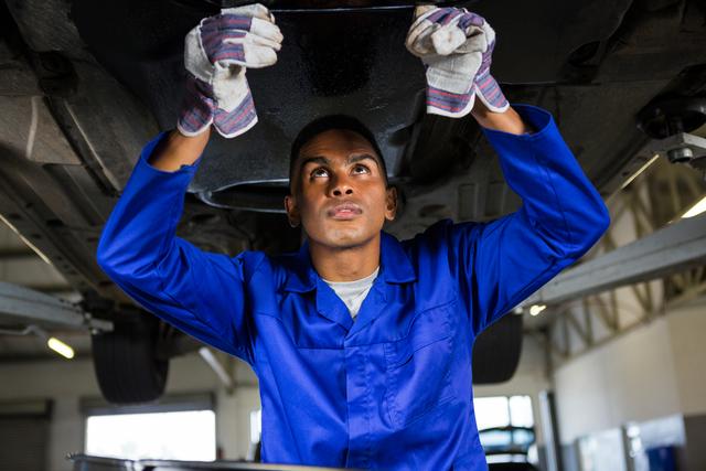 Mechanic wearing blue uniform and gloves working on car's undercarriage in a repair garage. Ideal for use in automotive industry promotions, service and maintenance advertisements, technical education materials, and mechanic training programs.