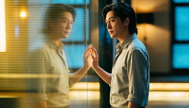 Asian man contemplating by glass partition pressing palm to reflection in moody office