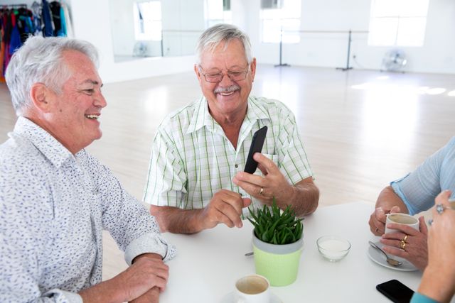 Group of happy senior male and female friends sitting at table together using smartphone socialising before ballroom dancing class, talking and drinking tea and coffee