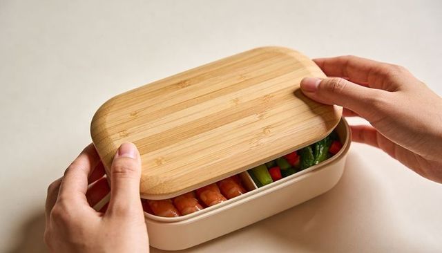 Lifting bamboo lid revealing bento lunchbox with shrimp and vegetables on beige tabletop