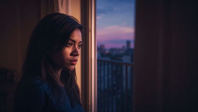 Contemplative young woman gazing through balcony glass at dusk skyline in urban apartment
