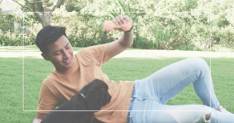 Asian man reclining on grass playing with black puppy holding pink bone toy outdoors