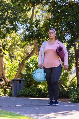 Plus-size woman walking to yoga in sunlit park carrying mat and duffel in urban activewear