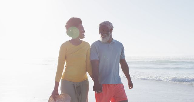 This image features a happy senior African American couple holding hands and walking along a picturesque beach. Ideal for promoting retirement, healthy living, travel destinations, and romantic getaways. It can be used in advertisements for beach resorts, retirement planning services, health magazines, and couples' counseling websites.