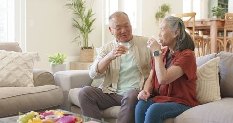 Asian senior couple sitting on sofa sipping water and smiling in bright modern living room