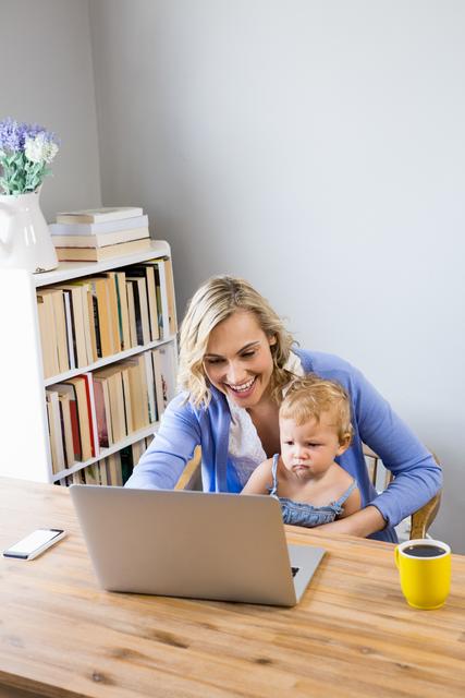 A mother wearing a blue sweater is working on a laptop while holding her baby on her lap. They are sitting at a wooden table in a cozy home environment with bookshelves and a cup of coffee nearby. This image can be used to depict themes of work-life balance, remote work, parenting, and family bond. Ideal for articles, blog posts, and advertisements related to parenting, home office setup, and family life.