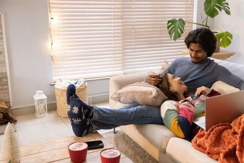 Diverse couple relaxing on cozy sofa with laptop, woman wearing striped knit sweater