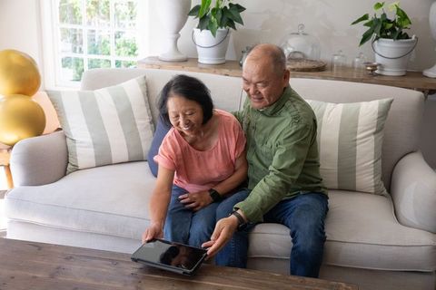 Asian senior couple sharing tablet on sofa in sunlit living room, cozy home tech