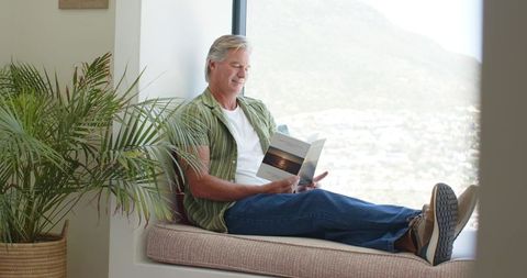 Mature man relaxing on window seat reading booklet with mountain view and sunlit interior