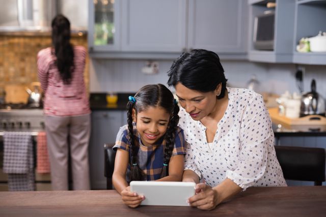 Grandmother and granddaughter using a digital tablet together in a cozy kitchen. Ideal for illustrating family bonding, technology use in everyday life, and multigenerational relationships. Perfect for advertisements, educational materials, and articles on family dynamics or technology in the home.