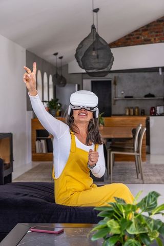Senior woman wearing vr headset reaching out while sitting on sofa in modern living room
