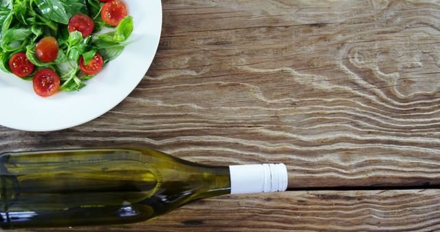 Image of a fresh salad with cherry tomatoes and greens next to a wine bottle on a wooden table. Useful for articles, menus, food blogs, lifestyle websites, and restaurant advertisements promoting healthy eating, dining experiences, or rustic meal presentations.