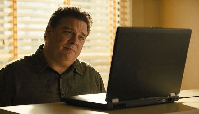 Middle-aged man working remotely on laptop at home with sunlit blinds and thoughtful expression