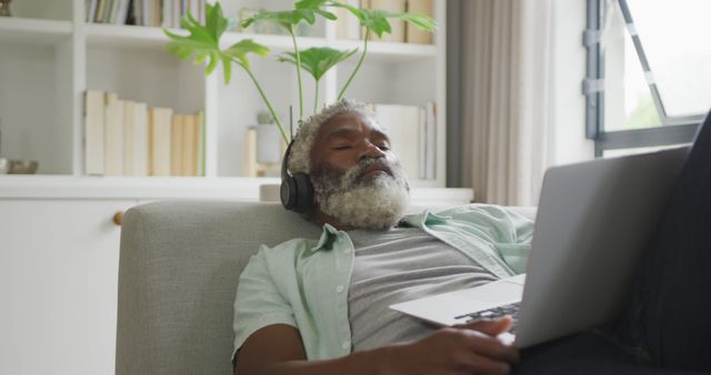 Senior man sitting on a couch while listening to music with headphones and using a laptop. Ideal for use in contexts related to technology use among seniors, relaxation, modern lifestyle, retirement activities, home comfort, digital literacy among the elderly, and living room decor.