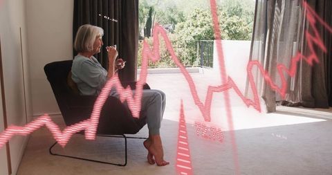 Senior woman relaxing in pajamas sipping tea by balcony with red market graph overlay