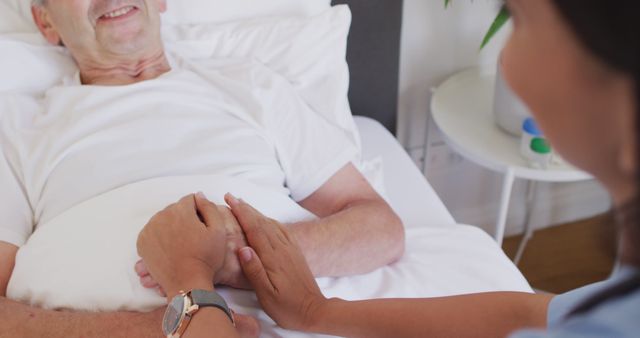 Elderly man resting in bed being comforted by caretaker holding his hands. This illustrates elderly care and the compassionate support provided by healthcare professionals. Useful for healthcare, nursing home, and patient care related content depicting trust, empathy, and the importance of compassionate care in medical settings.