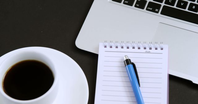 Image captures a work desk with a cup of coffee, a note pad with a pen, and an open laptop. Useful for illustrating themes around office work, productivity, freelancing, remote working, executive meetings, and business environments.