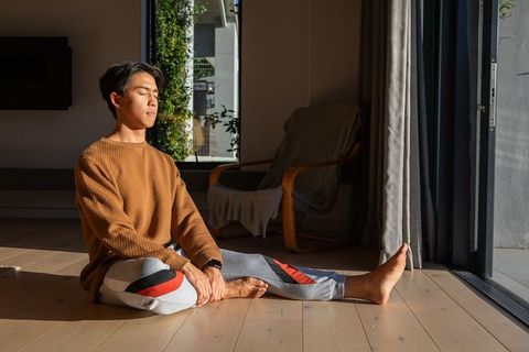 Asian man sitting on hardwood floor meditating in warm sunlight by sliding glass door