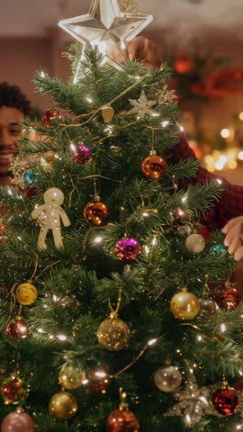 Couple placing star atop Christmas tree while decorating with glowing baubles and fairy lights
