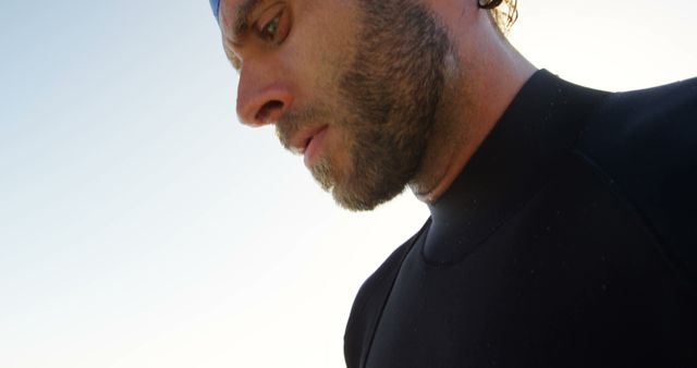This image shows a man wearing a wetsuit and swim cap, likely preparing for a swim at the beach. His serious expression suggests concentration or contemplation. This visual can be used in contexts related to sports, swimming, fitness, or advertisements promoting athletic gear, beach activities, or outdoor adventures.