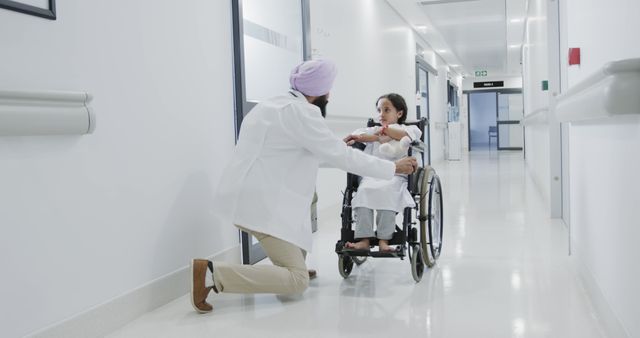 A doctor in white coat comfortting a child in a wheelchair in a hospital corridor. The child looks worried while the doctor kneels down to offer support. This image can be used for health care services, medical support for children, hospital environment, and doctor-patient interaction themes.