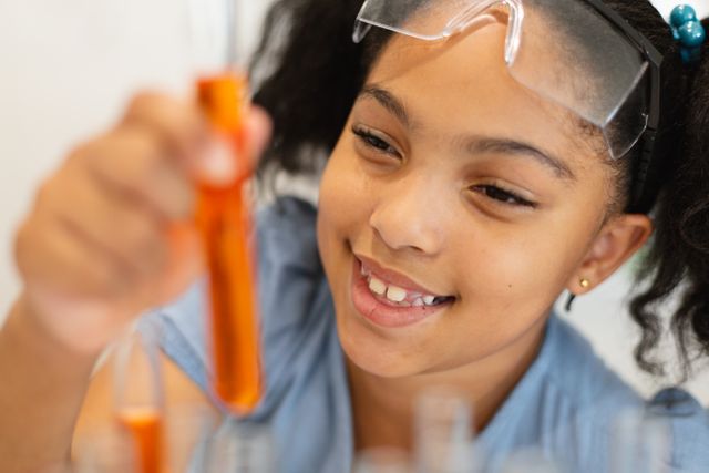 Young biracial schoolgirl wearing safety goggles, holding and examining a test tube filled with orange liquid during a chemistry class. Ideal for educational materials, STEM promotion, school science programs, and advertisements focusing on children's education and scientific curiosity.