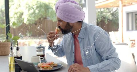 Indian man eating breakfast while using tablet at bright sunlit kitchen counter wearing turban