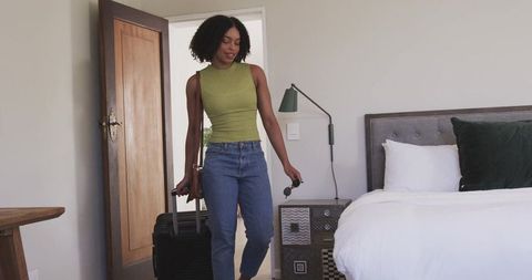 African american woman arriving in bedroom pulling suitcase holding sunglasses