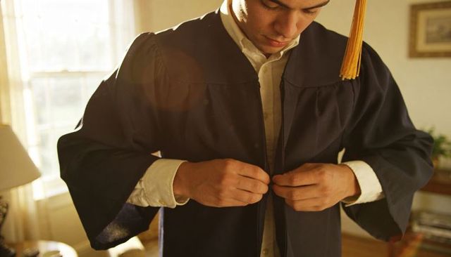 Young man buttoning graduation gown with gold tassel in sunlit living room ready for ceremony
