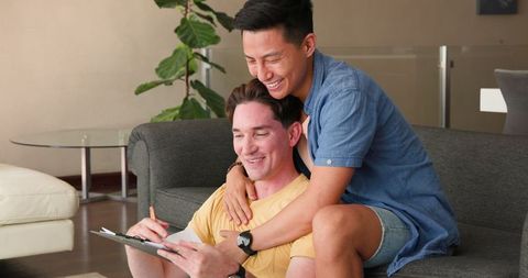 Smiling Diverse Gay Couple Reviewing Documents on Cozy Living Room Sofa