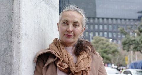Mature woman leaning on city column wearing tan coat and knit scarf, urban portrait