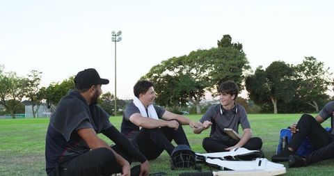 Youth cricket teammates sitting on field chatting and fist-bumping after practice