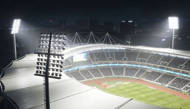 Floodlit night stadium hosting soccer match with packed crowd, players, track and skyline