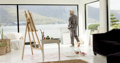 Indian male artist gazing at ocean from sunlit studio with easel and painting supplies