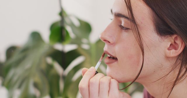 Young woman carefully applying lip gloss while surrounded by greenery. Ideal for beauty blogs, makeup tutorials, skincare advertisements, and lifestyle magazines highlighting personal care routines.