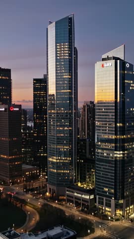 Vertical drone video at dusk revealing glass skyscrapers, illuminated roadways and plaza