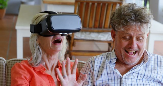 Senior couple sitting on a couch at home, enjoying virtual reality experience with a VR headset. The woman is wearing the headset and waving her hand, while the man is laughing and pointing. This image is perfect for use in articles about technology adoption among seniors, promoting family bonding through innovative tech, or illustrating the joy of new experiences at any age.