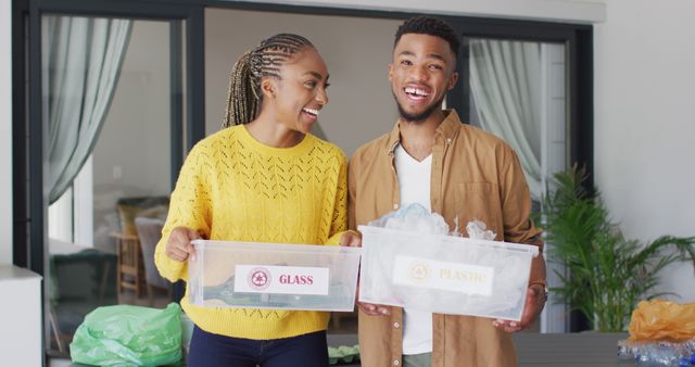 Young couple smiling while organizing recyclable materials in clear bins marked 'Glass' and 'Plastic'. Ideal for content on recycling, environmental awareness, sustainable living, and teamwork in the household.