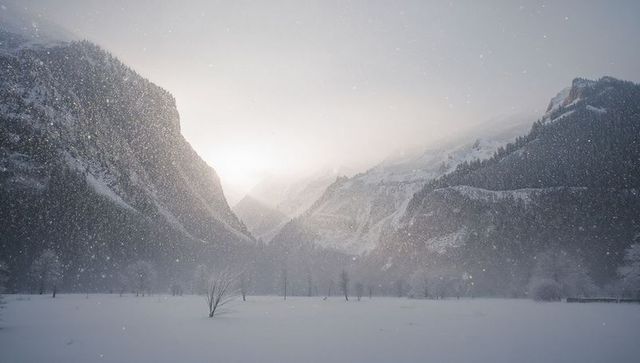 Sunlight glowing over snowy alpine valley with falling snow, misty mountain ridges, haze