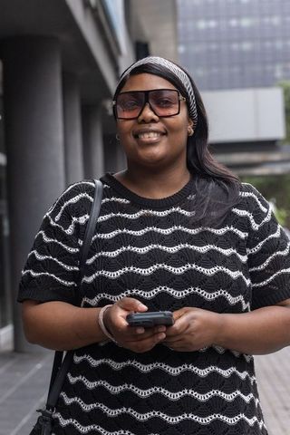 African american woman standing holding smartphone smiling wearing sunglasses knit top