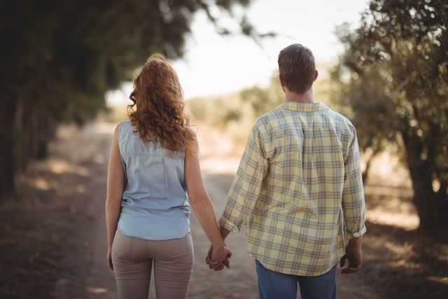 Rear view of couple holding hands while walking on dirt road at olive farm